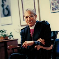 The Rt. Rev. Barbara Harris, sitting in a chair, smiling at the camera