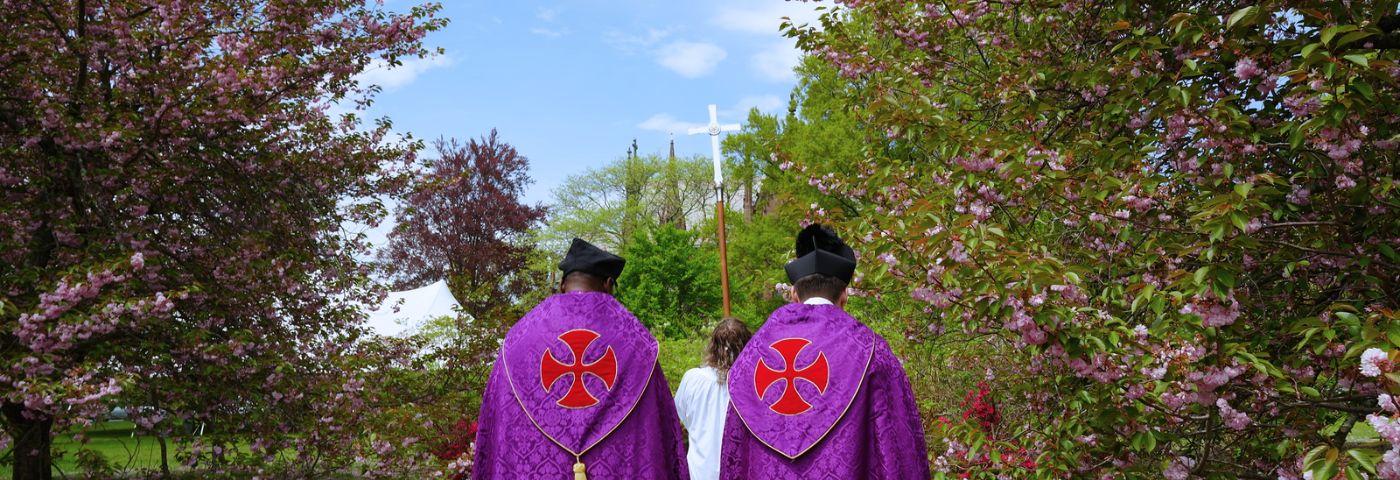 A view behind two priests in purple vestments following a processional cross through the grounds of the Cathedral of the Incarnation