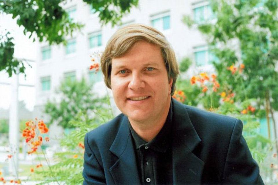 John Dear sitting in front of a blurred building with greenery around him
