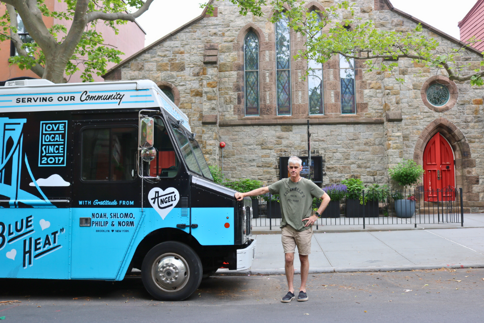 Fr. John Merz stands outside of a church next to the blue and black North Brooklyn Angels food truck